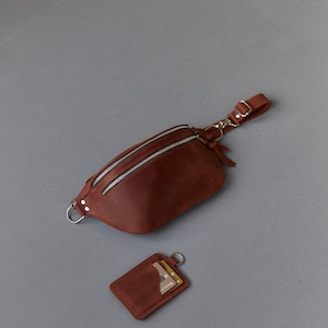 May include: A brown leather fanny pack with a zip closure and a detachable strap. A matching brown leather card holder with a silver ring is also included.