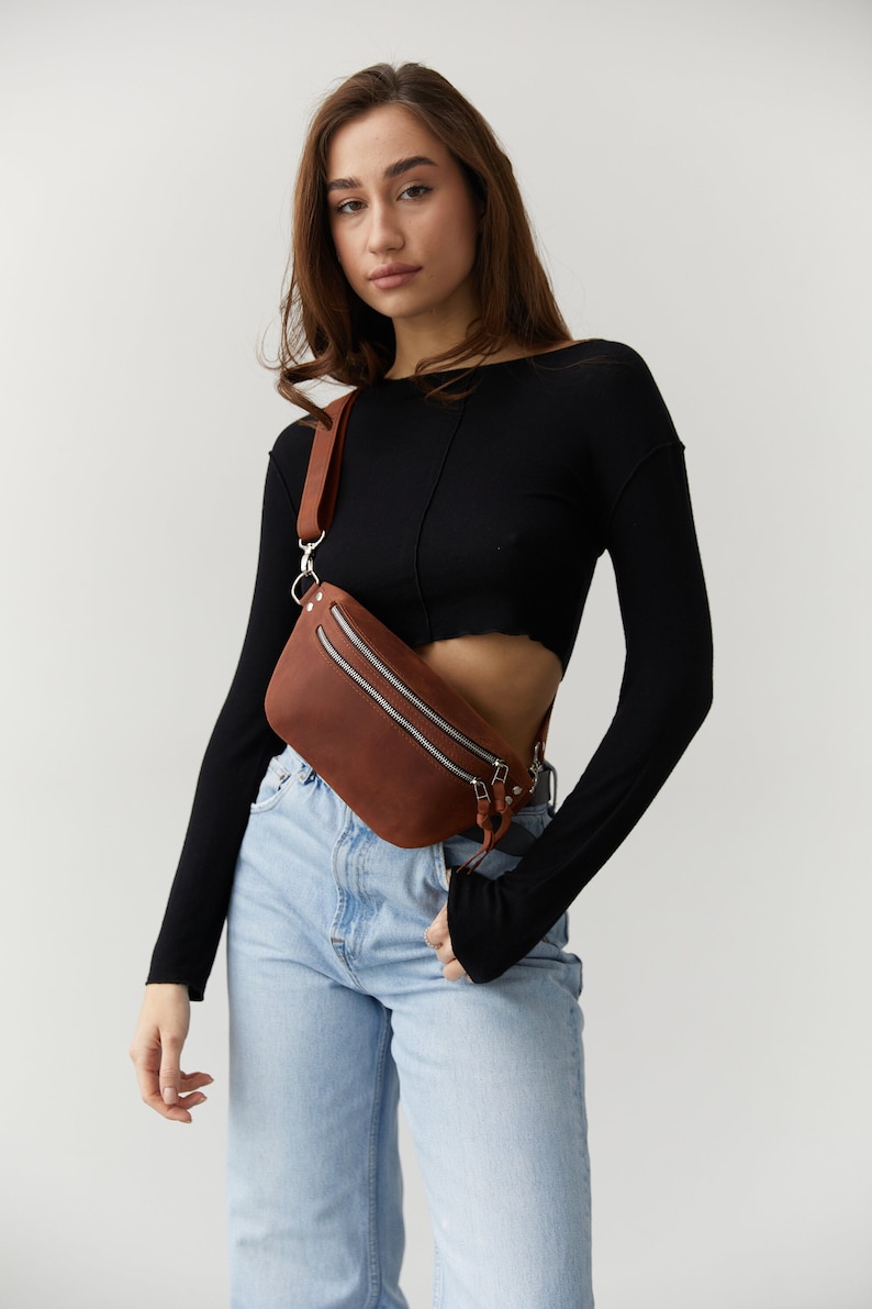 May include: A woman wearing a black long-sleeved shirt and light blue jeans with a brown leather fanny pack worn across her body.