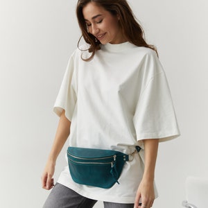 May include: A woman wearing a white t-shirt and grey jeans with a teal leather fanny pack.