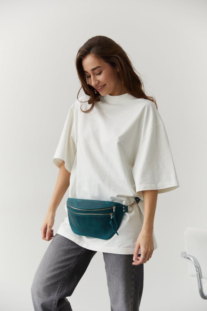 May include: A woman wearing a white t-shirt and grey jeans with a teal leather fanny pack.