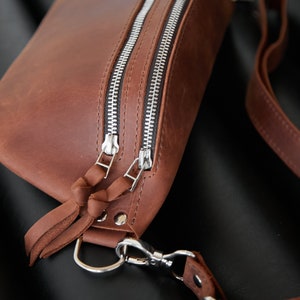 May include: A brown leather fanny pack with two silver zippers and a silver clasp. The bag has a long brown leather strap.