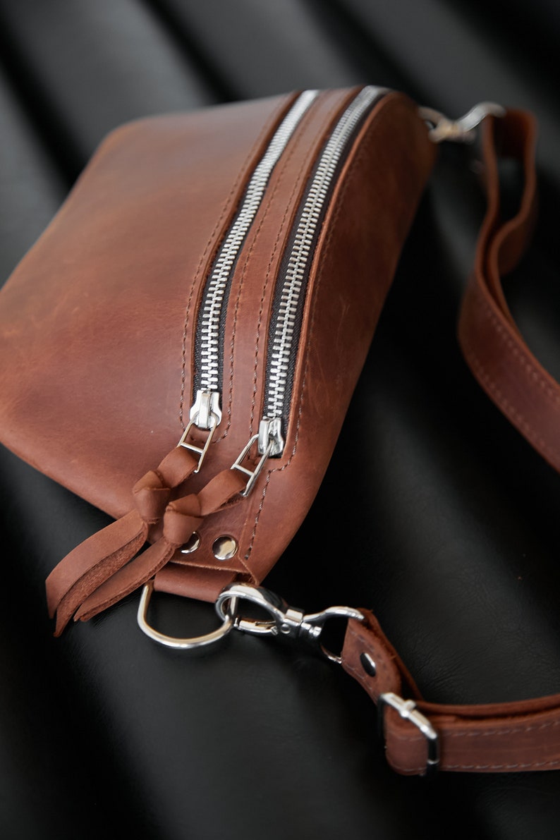 May include: A brown leather fanny pack with two silver zippers and a silver clasp. The bag has a long brown leather strap.