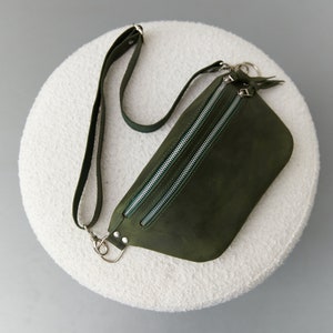 May include: A dark green leather fanny pack with two silver zippers and a silver strap. The bag is on a white fluffy surface.