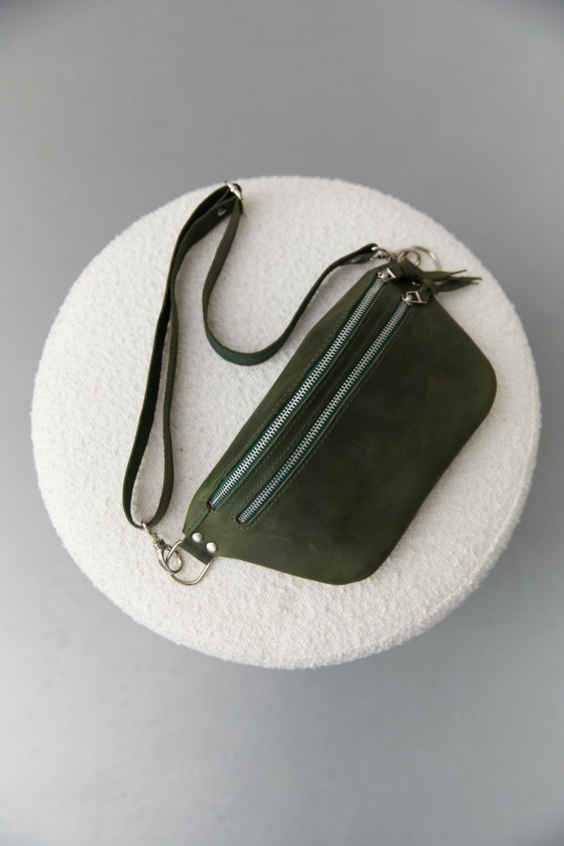May include: A dark green leather fanny pack with two silver zippers and a silver strap. The bag is on a white fluffy surface.