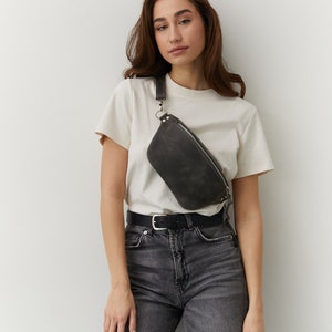 May include: A woman wearing a white t-shirt and gray jeans with a black leather fanny pack worn across her body.