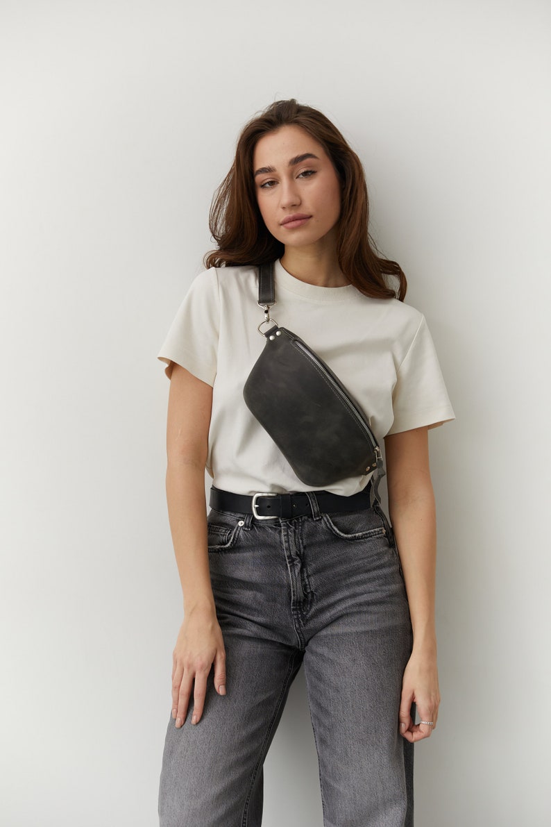 May include: A woman wearing a white t-shirt and gray jeans with a black leather fanny pack worn across her body.