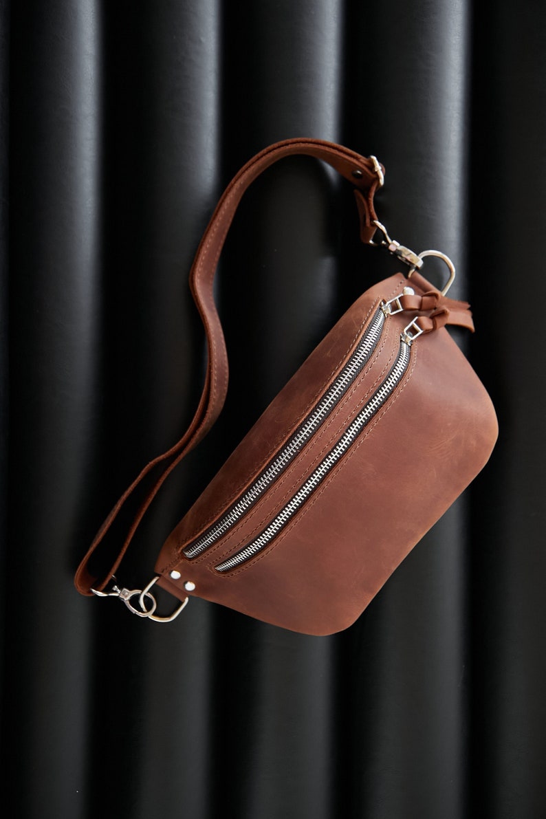 May include: A brown leather fanny pack with two silver zippers and a silver clasp. The bag is hanging on a black textured background.