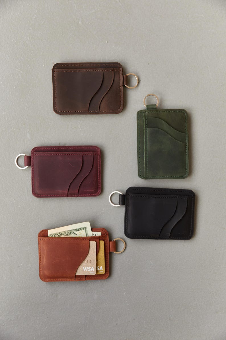 May include: Five leather wallets with key rings. The wallets are in brown, burgundy, green, black, and orange. Each wallet has a unique design with multiple pockets and a key ring.