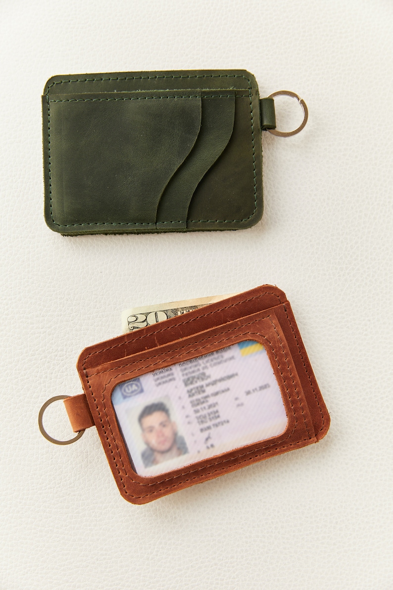 May include: Two leather wallets with key rings. The top wallet is dark green and the bottom wallet is brown with a clear window for an ID card. The brown wallet has a billfold and a US dollar bill is visible.