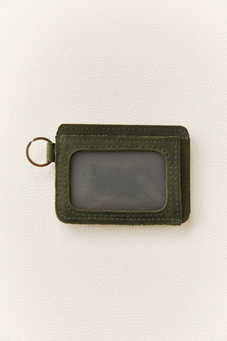May include: A dark green leather wallet with a clear plastic window for an ID card. The wallet has a metal ring attached to the side.