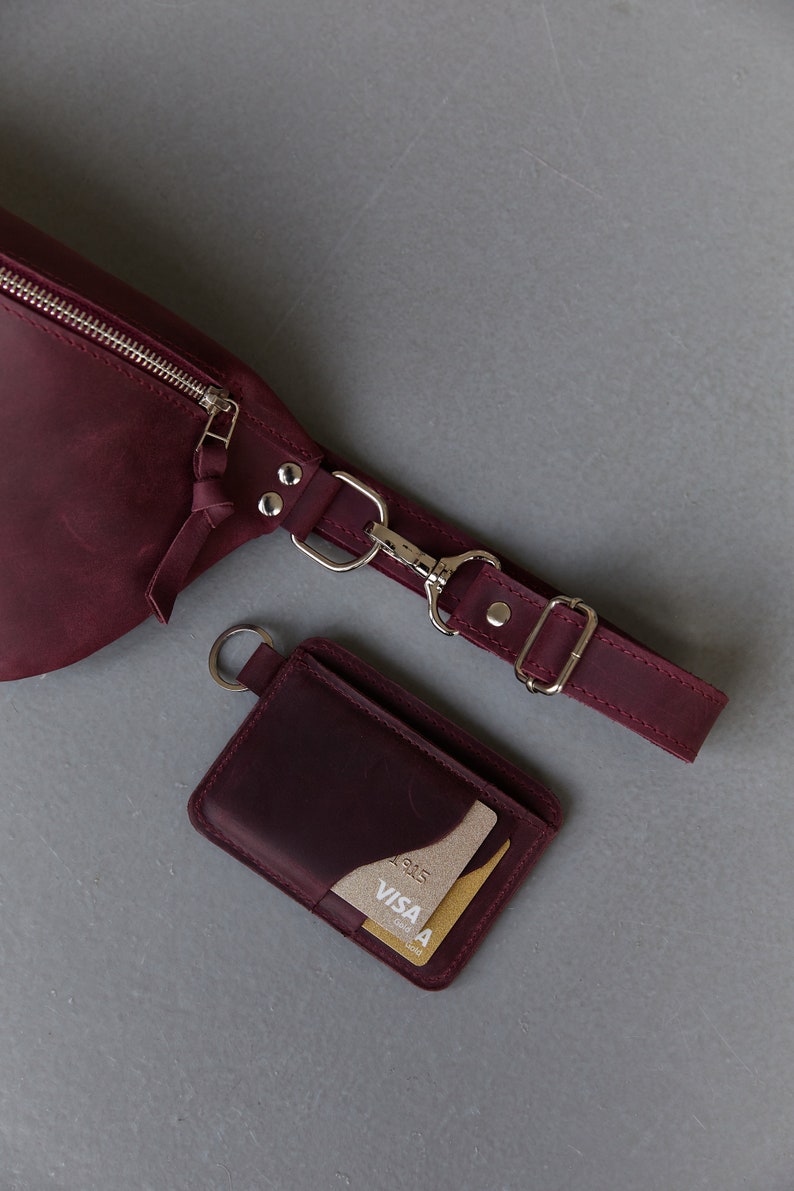May include: A burgundy leather fanny pack with a silver zip and a matching burgundy leather card holder with a gold VISA card inside.