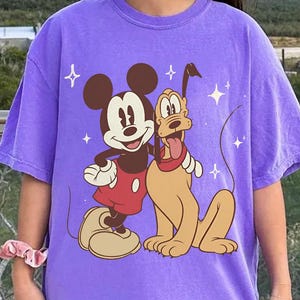 May include: A lilac t-shirt with a graphic of Mickey Mouse and Pluto. Mickey is in red shorts and Pluto has a red collar. The design includes star accents. The shirt is a casual, oversized style.