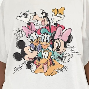 May include: White t-shirt featuring a colorful graphic of Disney characters including Mickey Mouse, Minnie Mouse, Donald Duck, Daisy Duck, Goofy, and Pluto. The shirt has short sleeves and a crew neck. The characters' names are written in cursive.