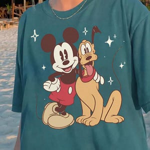 May include: Teal t-shirt with a graphic of Mickey Mouse and Pluto. Mickey wears red shorts, and Pluto has a red collar. The design includes white stars. The tee is made of a soft, comfortable material.
