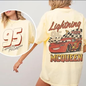 May include: A light yellow t-shirt featuring the number 95 and the words "Lightning McQueen" in a red and white design on the front. The back of the shirt displays a graphic of Lightning McQueen with the words "Lightning McQueen" and "Cars".