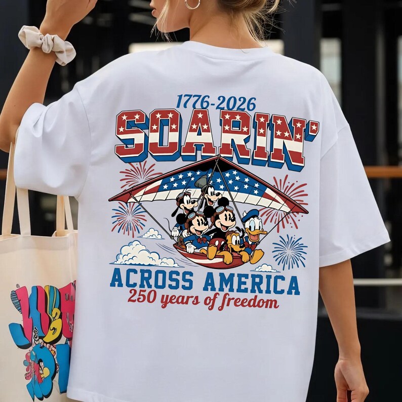 K&ouml;nnte beinhalten: Ein wei&szlig;es T-Shirt mit einer Grafik von Disney-Figuren in einem H&auml;ngegleiter im US-Flaggen-Design. Der Text lautet 1776-2026 SOARIN ACROSS AMERICA 250 years of freedom, umgeben von roten und blauen Feuerwerksk&ouml;rpern und Wolken.