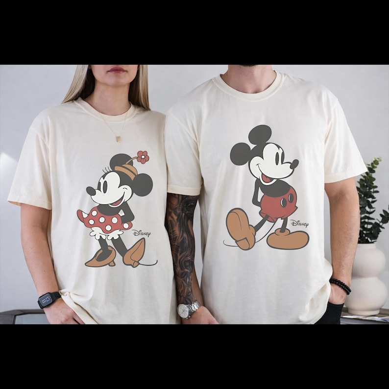 May include: Cream-colored t-shirts featuring Mickey and Minnie Mouse. Minnie is wearing a red polka-dot skirt and a flower on her head. Mickey is wearing red shorts and brown shoes. Both shirts have the Disney logo.