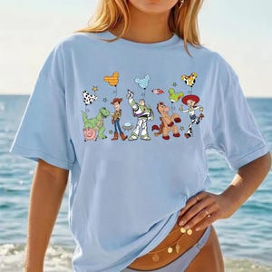 May include: Light blue t-shirt featuring a colorful graphic of animated characters and balloons. The design includes a dinosaur, cowboy, space ranger, horse, and cowgirl. The balloons have various patterns and shapes. The shirt is made of a soft material.