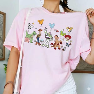 May include: A light pink t-shirt featuring a colorful graphic of Toy Story characters holding balloons. The design includes characters like Buzz Lightyear, Woody, and Jessie, with star and balloon accents. The shirt is a casual, short-sleeved style.