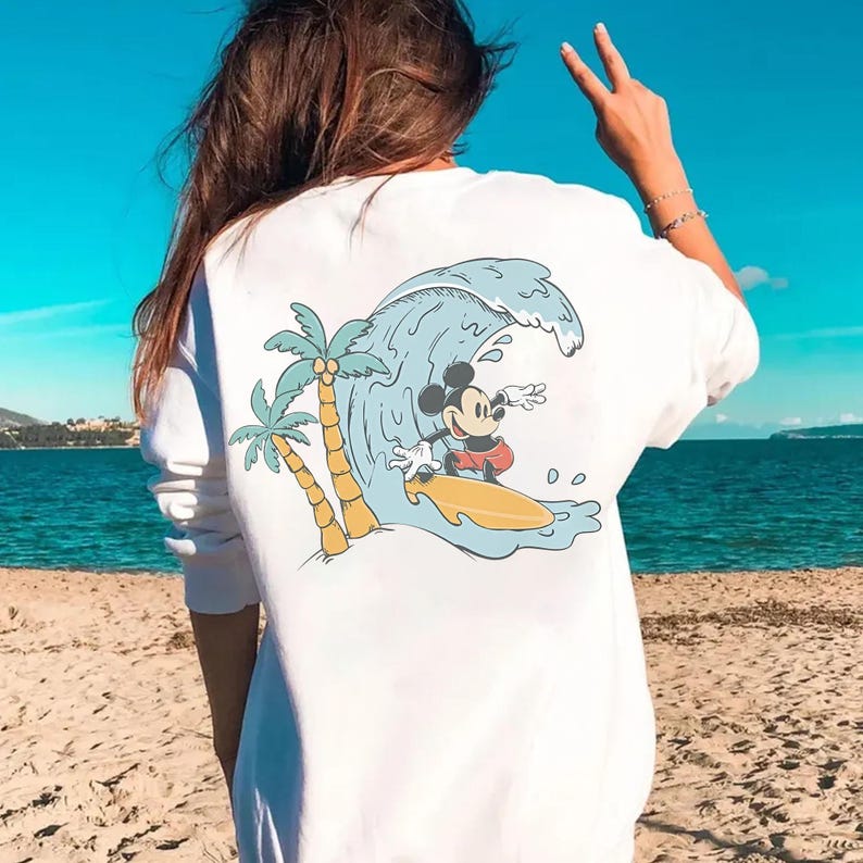 May include: White long-sleeve shirt featuring a cartoon graphic of Mickey Mouse surfing a wave. The design includes palm trees and a yellow surfboard. The background shows a beach and ocean under a bright blue sky.