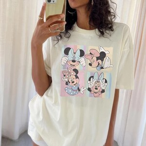 May include: Cream-colored t-shirt featuring four panels with Minnie Mouse illustrations. Each panel showcases a different pose of Minnie Mouse, with colorful backgrounds. The shirt has short sleeves and a relaxed fit, suitable for casual wear.