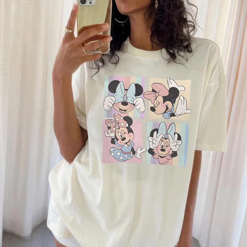 May include: Cream-colored t-shirt featuring four panels with Minnie Mouse illustrations. Each panel showcases a different pose of Minnie Mouse, with colorful backgrounds. The shirt has short sleeves and a relaxed fit, suitable for casual wear.