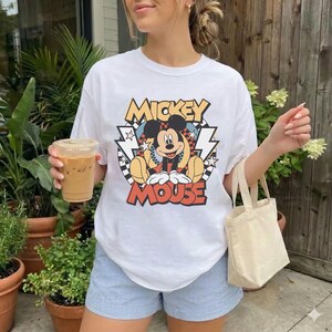May include: White t-shirt featuring a colorful graphic of Mickey Mouse with the text "Mickey Mouse". The design includes yellow, red, black, and blue elements. The shirt is paired with light blue shorts.