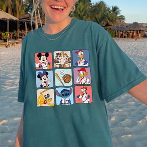 May include: Teal-colored t-shirt featuring a grid of cartoon characters in baseball-themed poses. Mickey Mouse, Minnie Mouse, Donald Duck, and Goofy are among the characters. The shirt has a relaxed fit and short sleeves.