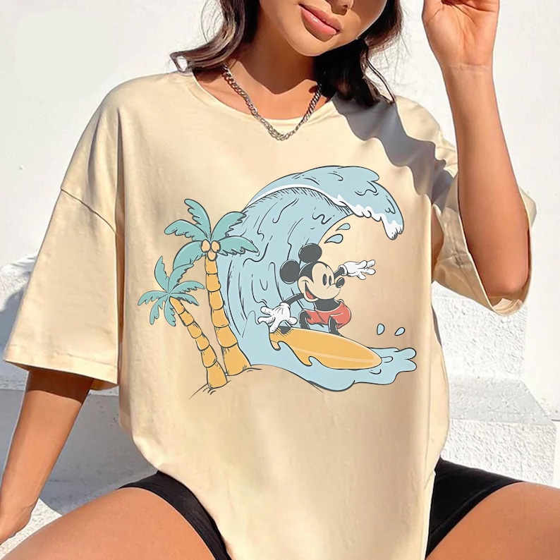 May include: Cream-colored t-shirt featuring a cartoon of Mickey Mouse surfing a wave. The design includes palm trees and a blue wave. The shirt has short sleeves and a relaxed fit, perfect for casual wear.
