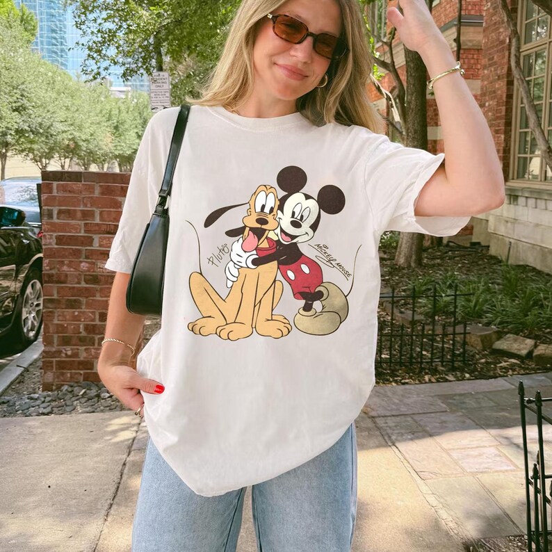 May include: Off-white t-shirt featuring a graphic of Mickey Mouse and Pluto embracing. The cartoon characters are in color, with Mickey in red shorts and Pluto in yellow. The shirt is paired with light blue jeans.