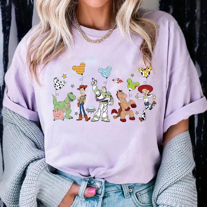 May include: A light purple t-shirt featuring a graphic of characters from the animated movie Toy Story. The design includes Woody, Buzz Lightyear, and other characters with colorful balloons. The shirt is made of a soft material.