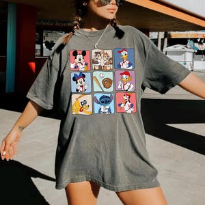 May include: Gray oversized t-shirt featuring a grid of cartoon characters in baseball attire. The grid includes Mickey Mouse, Minnie Mouse, Donald Duck, Chip and Dale, Stitch, and Goofy. The shirt is worn by a person.