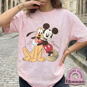 May include: A light pink t-shirt featuring a graphic of Mickey Mouse hugging Pluto. Mickey Mouse is wearing red shorts and Pluto is tan. The text "Mickey Mouse" and "Pluto" are also visible on the shirt.