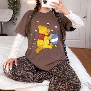 May include: A brown t-shirt featuring a cartoon illustration of Winnie the Pooh holding a honey pot, surrounded by bees. The shirt is paired with leopard print pants and a white long-sleeved shirt.