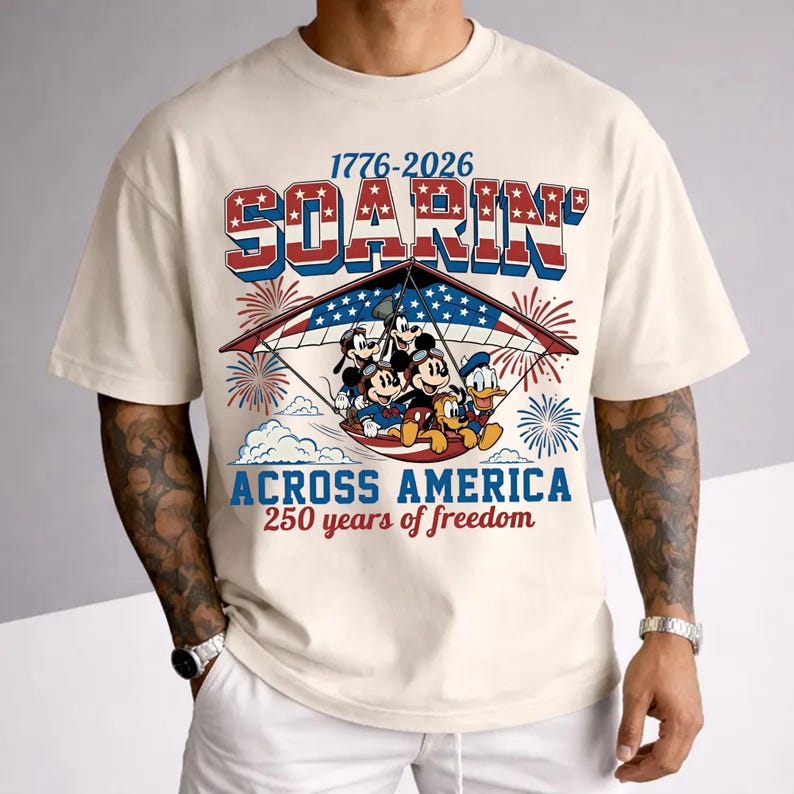 K&ouml;nnte beinhalten: Ein cremefarbenes T-Shirt mit einer Grafik von Disney-Figuren in einem H&auml;ngegleiter und dem Text 1776-2026 SOARIN' ACROSS AMERICA 250 years of freedom, akzentuiert durch rot-wei&szlig;-blaue Feuerwerke und ein Sternenbanner-Muster.