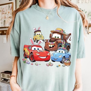 Può includere: T-shirt azzurra con una grafica a cartoni animati di quattro personaggi di Cars decorati per Pasqua. Le auto, tra cui Saetta McQueen e Cricchetto, sono decorate con uova e fiori pasquali.