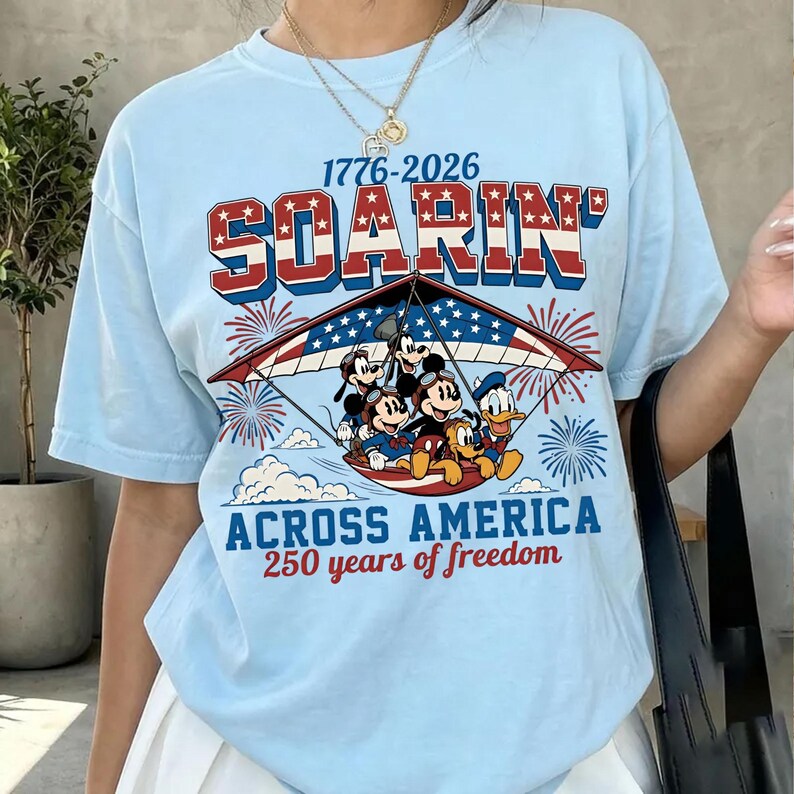 K&ouml;nnte beinhalten: Ein hellblaues T-Shirt mit einer Grafik von Disney-Figuren, die einen H&auml;ngegleiter im US-Flaggen-Design fliegen. Der Text lautet 1776-2026 SOARIN ACROSS AMERICA 250 years of freedom, erg&auml;nzt durch rote und blaue Feuerwerksdarstellungen.