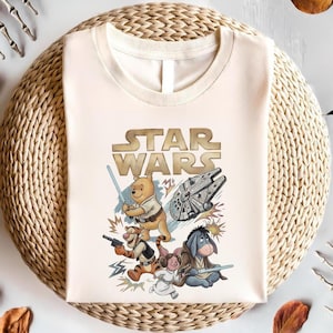 Könnte beinhalten: Cremefarbenes T-Shirt mit einem Mashup aus Star Wars und Winnie-the-Pooh-Charakteren. Das Design zeigt das Star Wars-Logo, Winnie Puuh mit einem Lichtschwert, Tigger mit einem Blaster und I-Aah mit einem Lichtschwert.