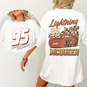 May include: White t-shirt featuring a graphic of Lightning McQueen from the Cars movie. The front displays the number "95" with "Lightning McQueen" below. The back showcases McQueen with a checkered flag and the word "MCQUEEN".