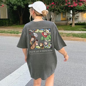 May include: Dark gray oversized t-shirt featuring a colorful graphic of animated characters, including a cowboy, a space ranger, and aliens. The text "YOU'VE GOT A FRIEND IN ME" is printed below the graphic. The shirt is worn casually.