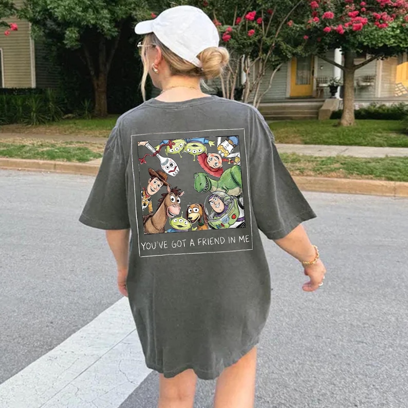May include: Dark gray oversized t-shirt featuring a colorful graphic of animated characters, including a cowboy, a space ranger, and aliens. The text "YOU'VE GOT A FRIEND IN ME" is printed below the graphic. The shirt is worn casually.
