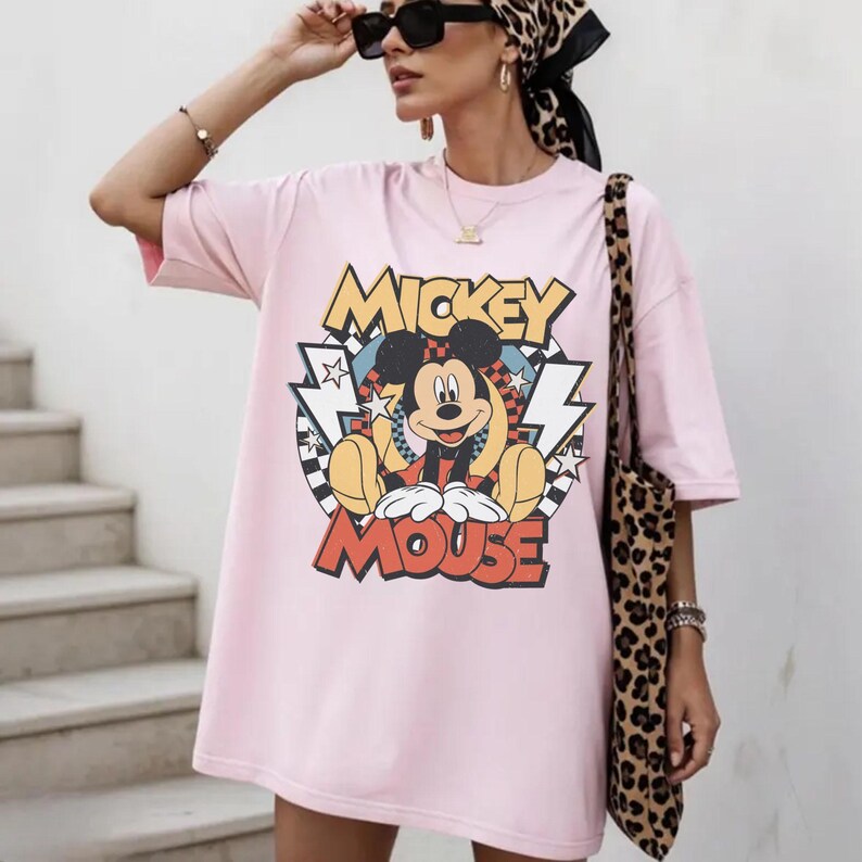 May include: A light pink oversized t-shirt featuring a graphic of Mickey Mouse. The design includes the words "Mickey Mouse" in yellow and red, with a cartoon image of Mickey. The shirt is paired with a leopard print tote bag.