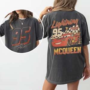 May include: Dark gray t-shirt featuring a large "95" graphic in red and orange on the front, with "Lightning McQueen" text. The back displays a red cartoon race car with "Lightning McQueen" and "95" text, and a checkered flag.