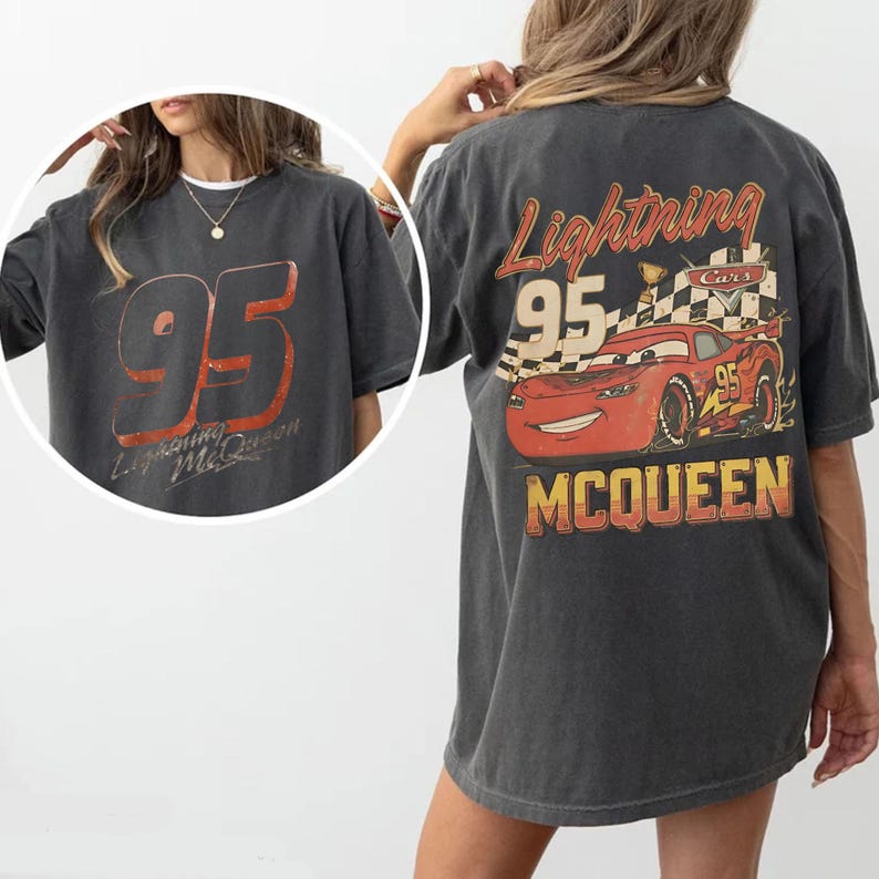 May include: Dark gray t-shirt featuring a large "95" graphic in red and orange on the front, with "Lightning McQueen" text. The back displays a red cartoon race car with "Lightning McQueen" and "95" text, and a checkered flag.