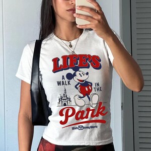 Puede incluir: Camiseta blanca con el texto "LIFE'S A WALK IN THE PARK" en rojo y azul, con Mickey Mouse y un gr&aacute;fico de castillo. La camiseta tambi&eacute;n incluye el texto "Walt Disney World".