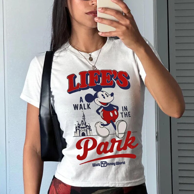 Puede incluir: Camiseta blanca con el texto "LIFE'S A WALK IN THE PARK" en rojo y azul, con Mickey Mouse y un gr&aacute;fico de castillo. La camiseta tambi&eacute;n incluye el texto "Walt Disney World".