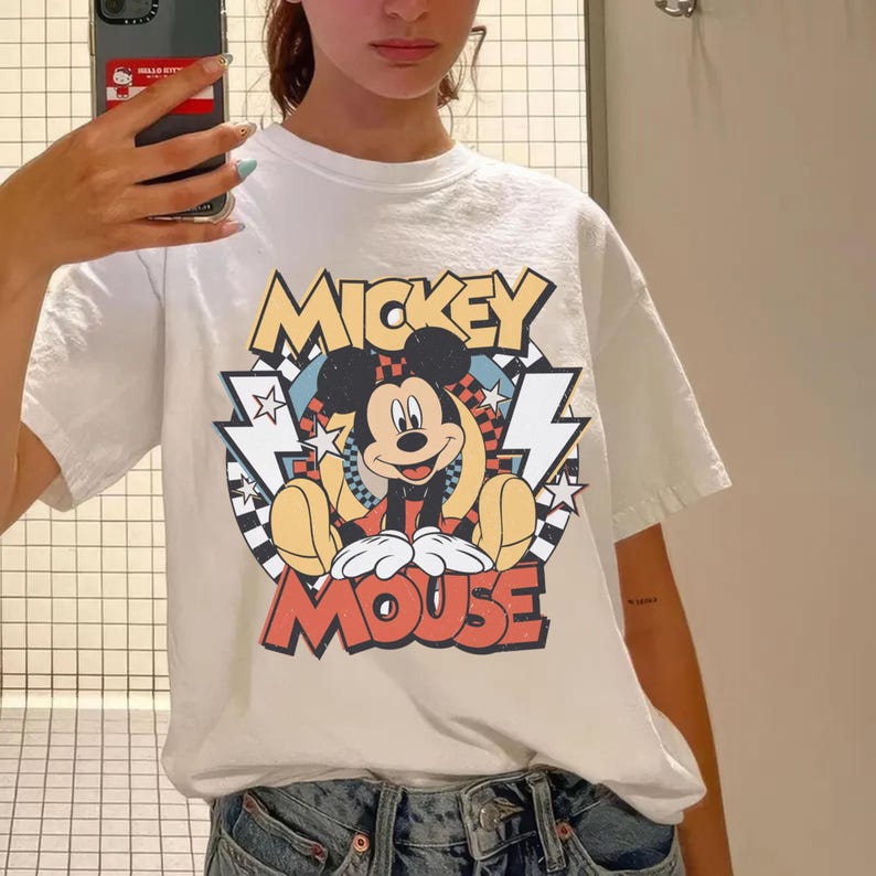 May include: White t-shirt featuring a colorful graphic of Mickey Mouse. The design includes the words "Mickey Mouse" in a retro font, with Mickey sitting in the center. The shirt has short sleeves and a crew neck.