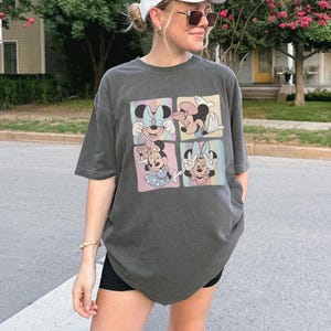 May include: Dark gray t-shirt featuring a four-panel graphic of Minnie Mouse in various poses. Each panel has a pastel rainbow background. The shirt is a relaxed fit and has short sleeves.