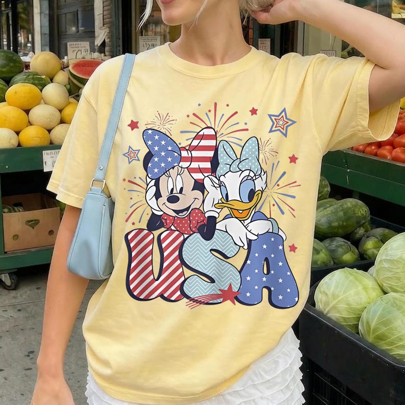 May include: Yellow t-shirt featuring Minnie Mouse and Daisy Duck with a patriotic "USA" design. The graphic includes red, white, and blue elements, stars, and fireworks. The shirt has short sleeves and a crew neck.