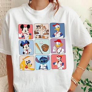 May include: White t-shirt featuring a grid of cartoon characters in baseball uniforms. Mickey Mouse, Chip and Dale, Donald Duck, Minnie Mouse, a baseball, glove, Daisy Duck, Pluto, and Stitch are depicted in colorful squares.